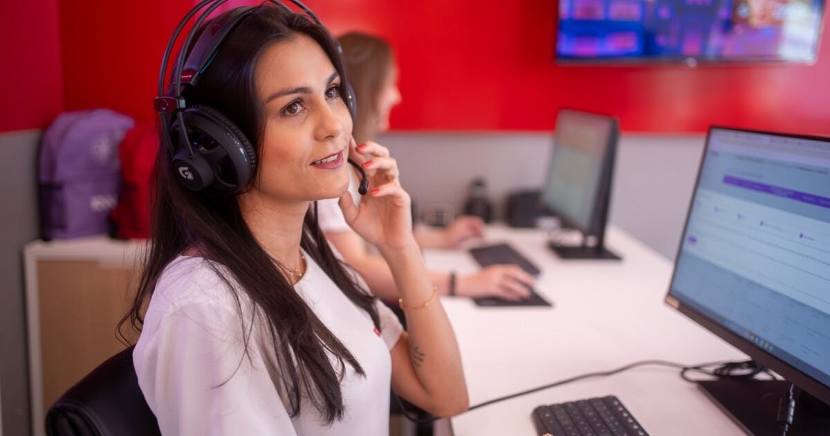 Mulher de camiseta branca, sentada, com um computador na frente e um fone de ouvido.