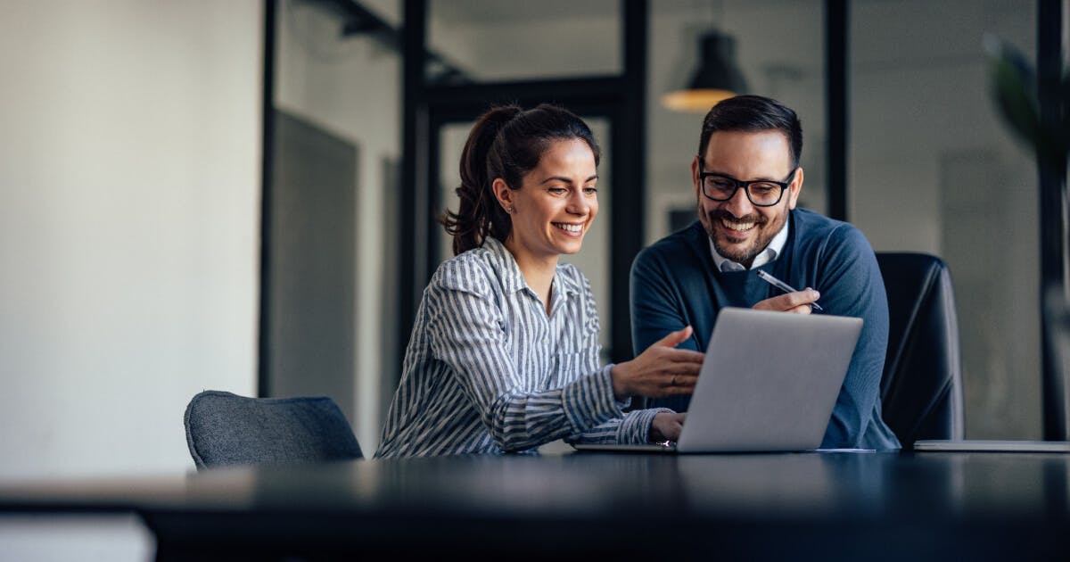 Homem e mulher sentados à mesa com um laptop, discutindo ou trabalhando juntos em um ambiente colaborativo.