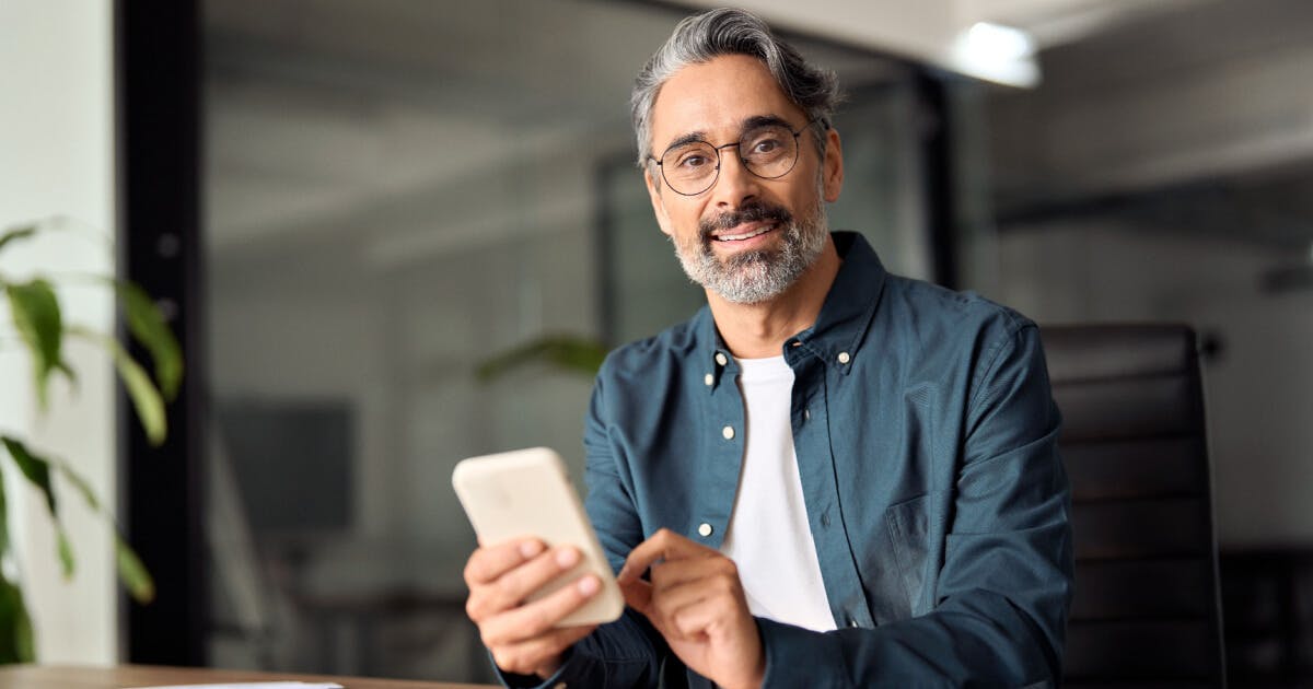 Homem de óculos e barba segurando um celular, com expressão concentrada e ambiente neutro ao fundo.