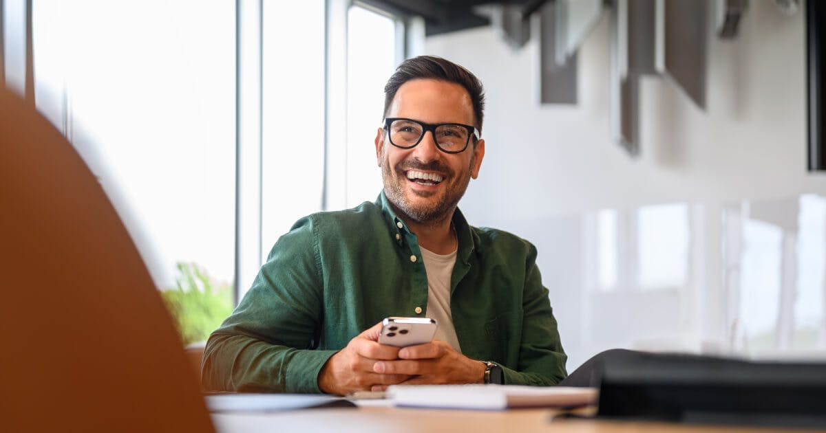 Homem de óculos sorrindo enquanto utiliza seu celular.