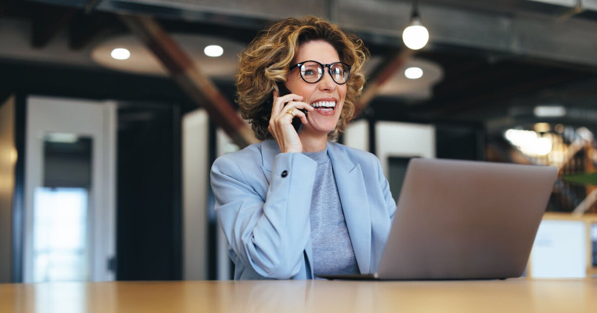 Uma mulher de óculos conversando ao telefone enquanto está sentada em uma mesa.