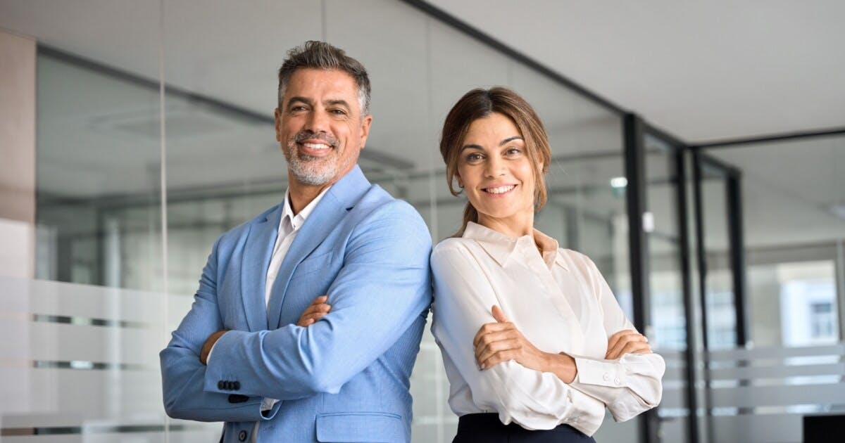 Uma mulher e um homem de braços cruzados encostados um no outro e sorrindo para a foto.