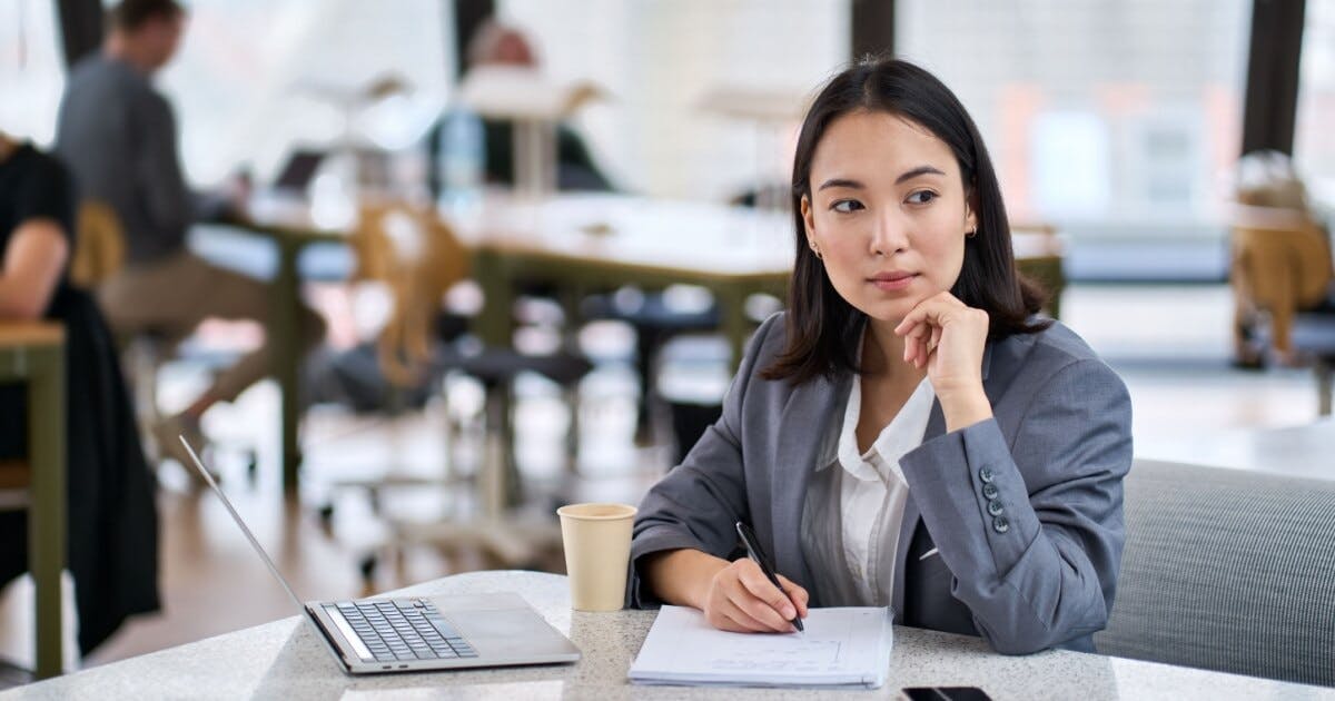 Uma mulher sentada em uma mesa com uma caneta na mão com expressão de dúvida. Tem um copo de café e um notebook na mesa.