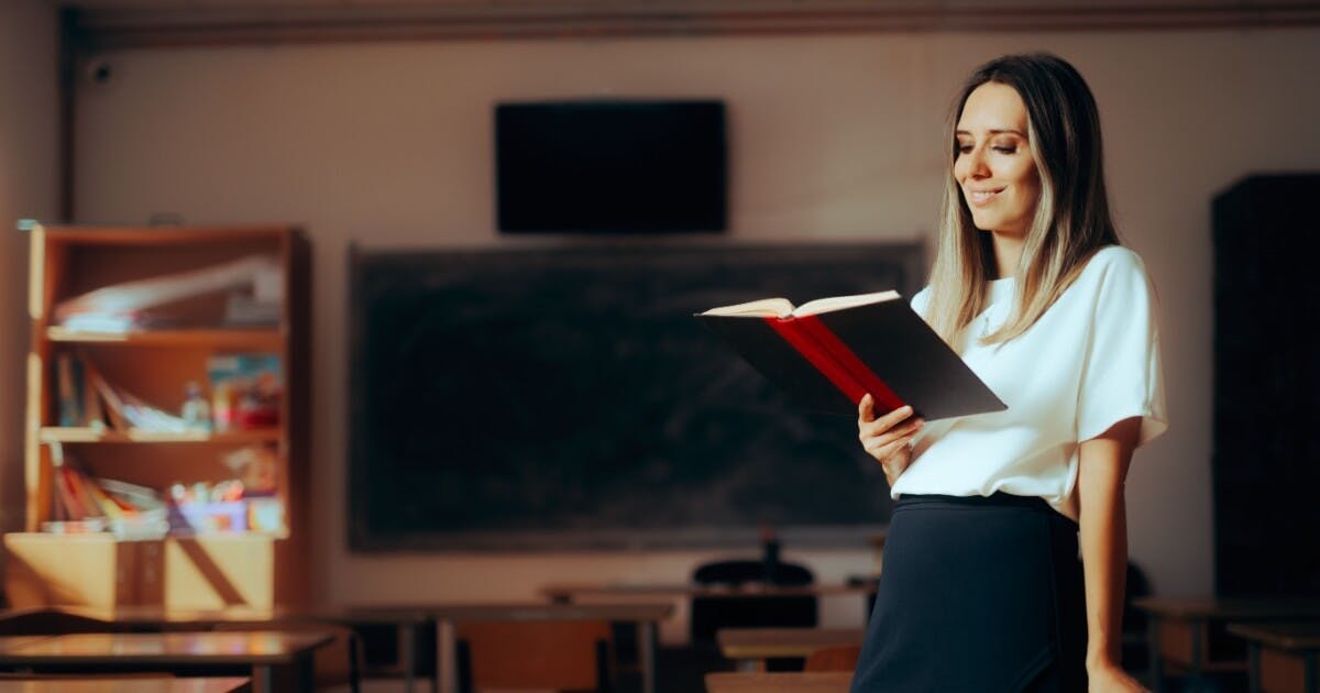 Professora sozinha em uma sala de aula olhando para um livro e sorrindo.