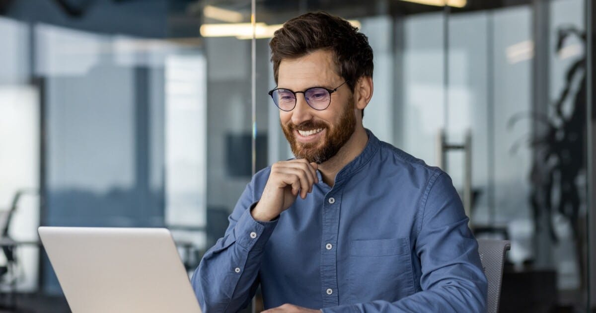 Homem de barba e óculos sorrindo, sentado e olhando para o notebook.