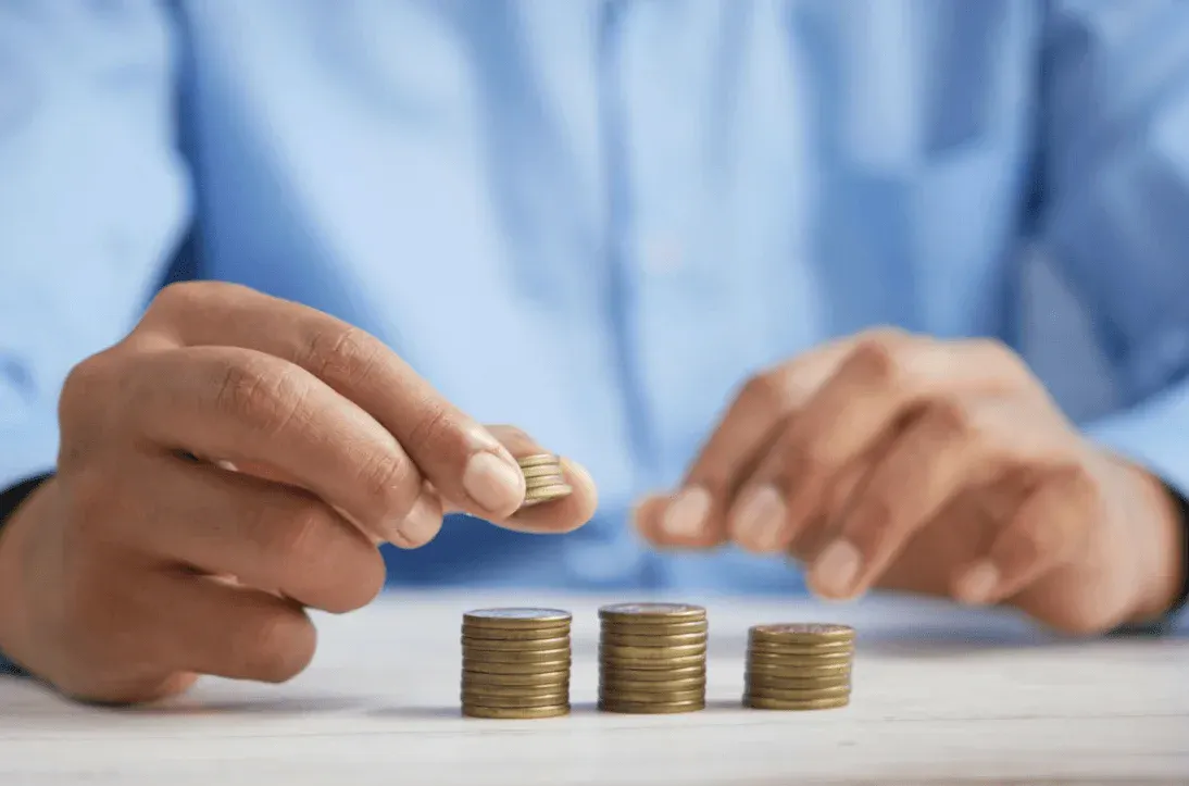 Pessoas em ambiente de escritório segurando uma moeda e colocando-a sobre uma pilha de moedas, sugerindo economia, investimento e crescimento financeiro.
