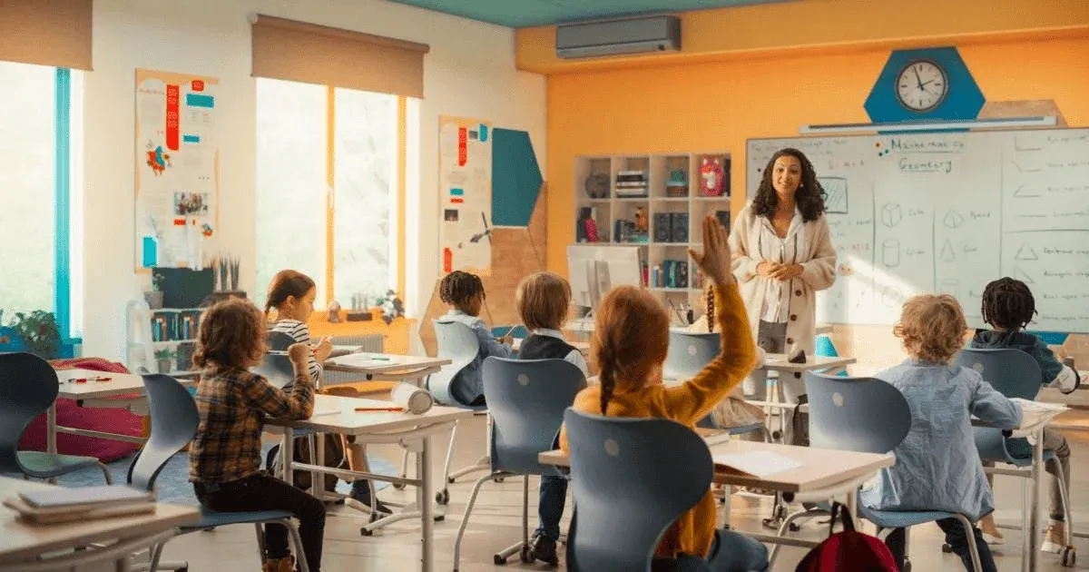 Sala de aula com crianças sentadas em mesas e professora em pé, apontando para um quadro branco com anotações e materiais didáticos ao fundo