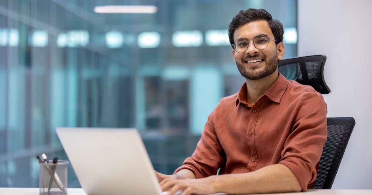 Homem de óculos sorrindo e com expressão de felicidade, sentado em uma mesa com um notebook em cima.
