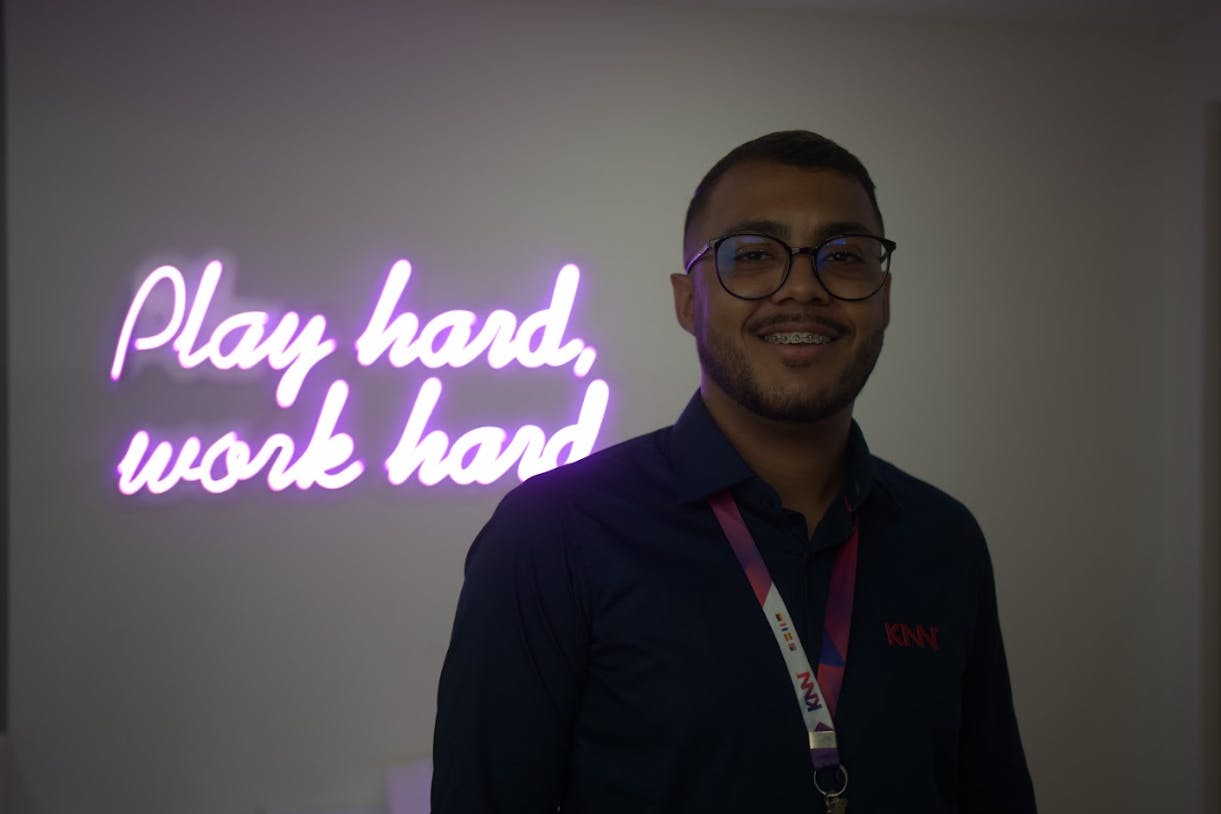 Homem de óculos, com o crachá da KNN Idiomas e sorrindo. Atrás, a frase "Play hard, work hard." em LED.