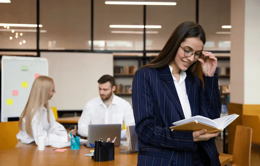 Mulher em escritório corporativo usando óculos e sorrindo enquanto lê um livro aberto, com colegas trabalhando ao fundo em mesas e computadores.