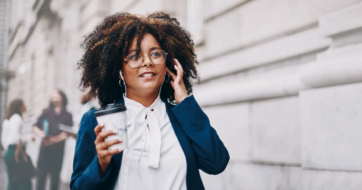 Mulher caminhando com um copo de café na mão, fones de ouvido e a mão esquerda no ouvido.
