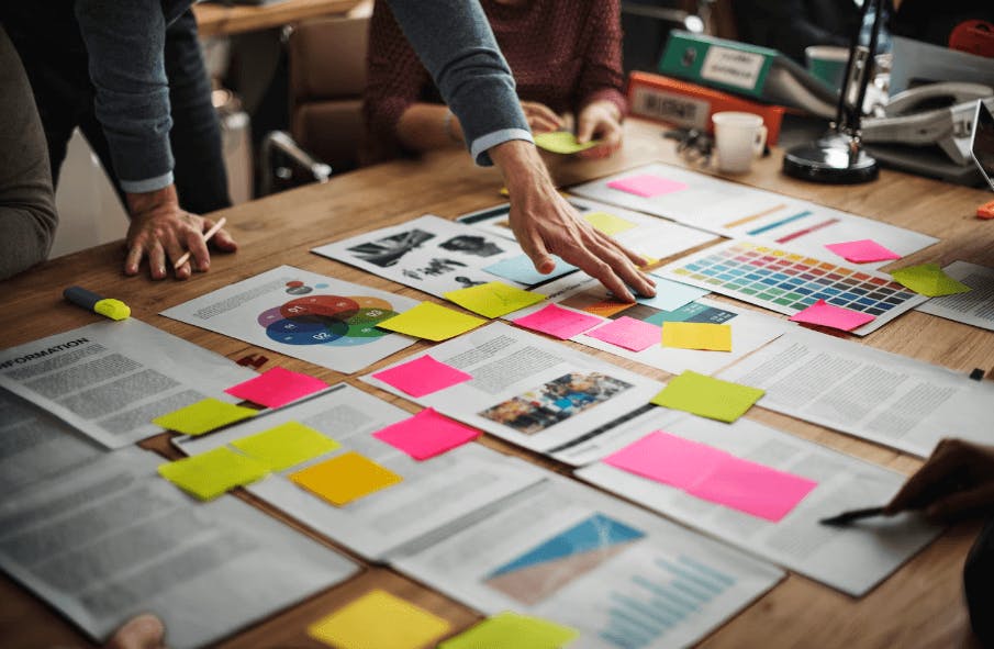 Equipe de profissionais planejando uma estratégia de marketing ou criando conteúdo para uma campanha, utilizando jornal, post-its coloridos e materiais de revisão.