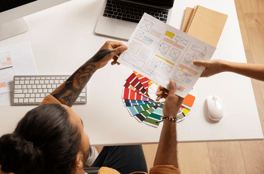 Designers discutindo paletas de cores e layout de projeto de design gráfico em escritório moderno, com computador portátil, papéis e caderno na mesa.