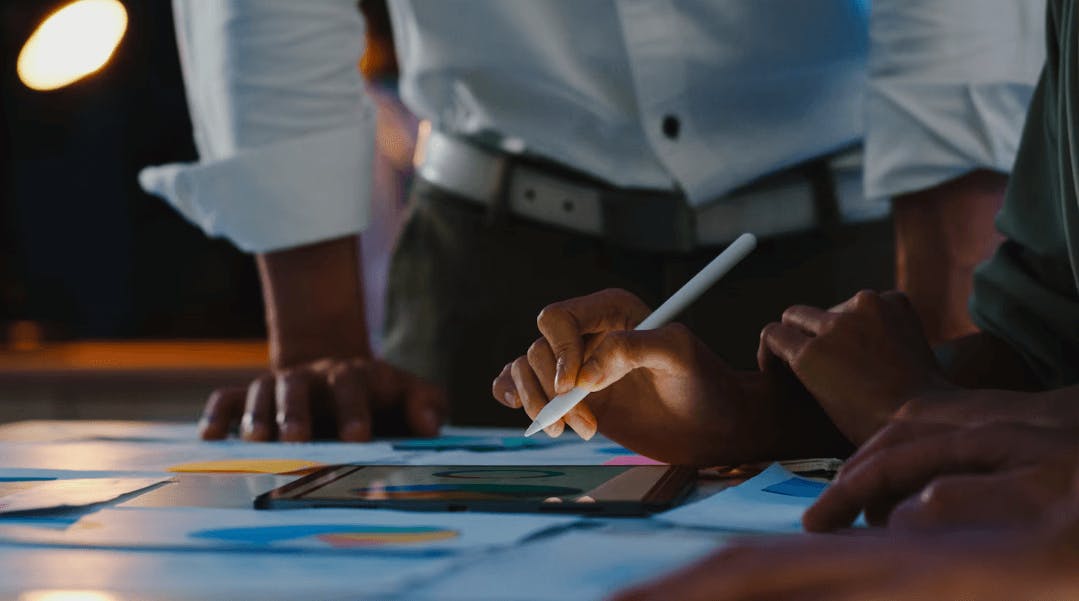 Pessoa usando um stylus para apresentar gráficos e dados em um tablet durante uma reunião de negócios, com outros participantes ao redor.