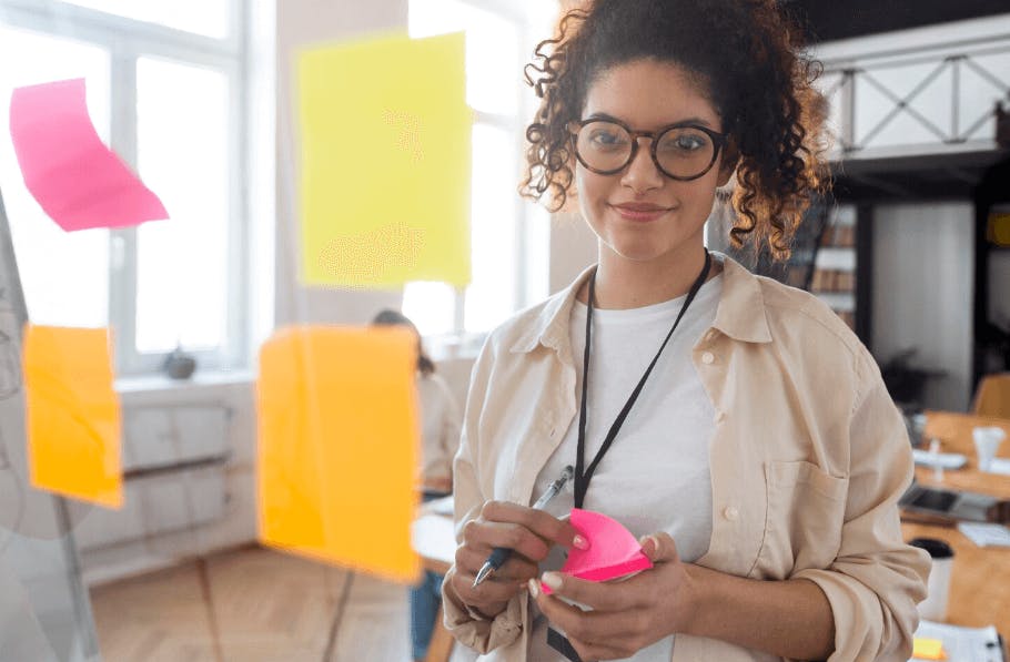 Mulher jovem de óculos com cabelo cacheado e sorriso, segurando um marcador rosa, em ambiente de escritório com post-its coloridos nas janelas ao fundo.
