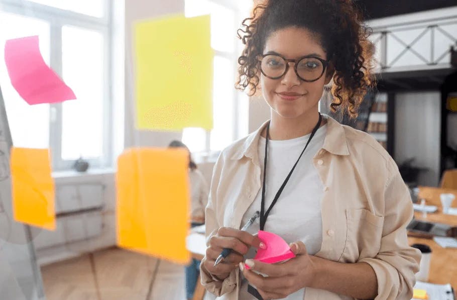 Mulher de óculos escuros e cabelo cacheado segurando post-it rosa em escritório criativo com parede de janelas e post-its coloridos ao fundo.