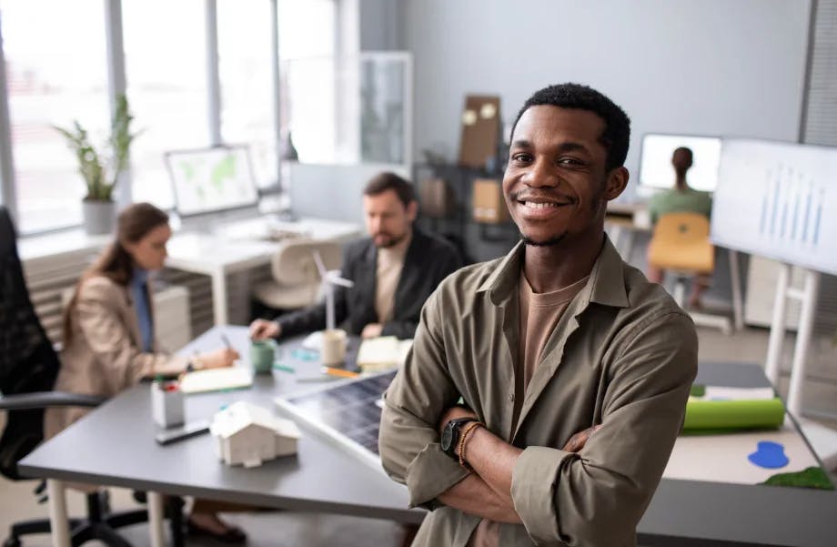 Homem sorridente na frente de uma mesa de reunião com colegas de trabalho em um ambiente de escritório moderno, com plantas e telas de apresentação ao fundo.