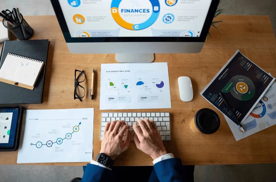Pessoa trabalhando em um escritório com múltiplos dispositivos eletrônicos, analisando gráficos financeiros e dados de finanças em computador, tablet e documentos.
