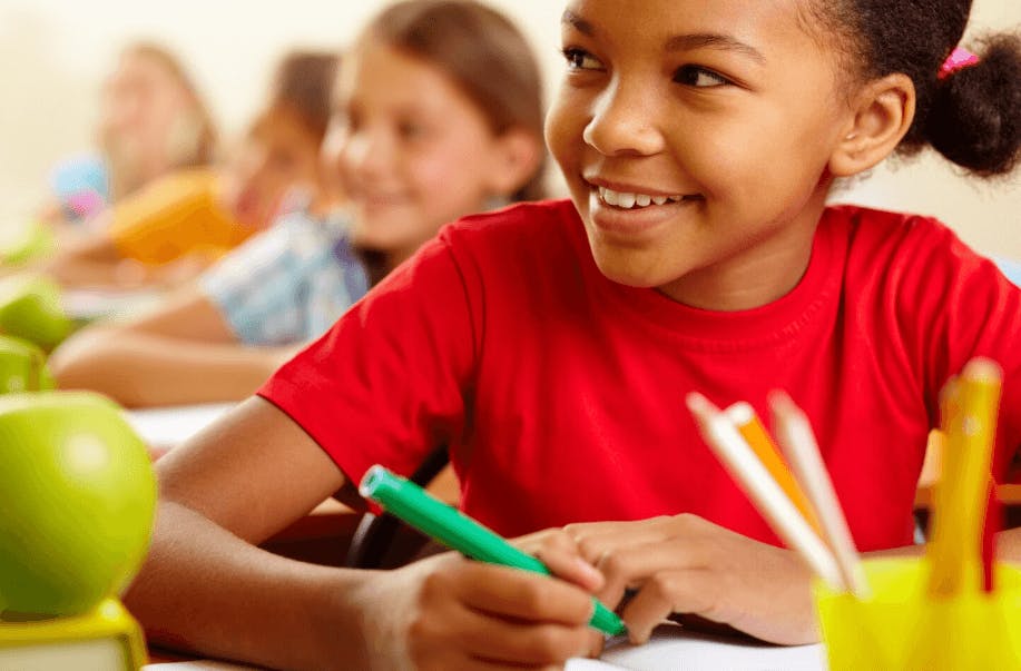 Criança sorrindo durante aula na escola, rodeada de colegas, com materiais escolares e maçã na mesa, simbolizando educação de qualidade e aprendizado.