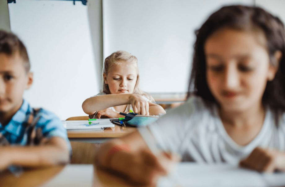Três crianças estudando em sala de aula com atenção, uma criança ajustando canetas e cadernos na mesa, ambiente escolar bem iluminado e direcionado para educação