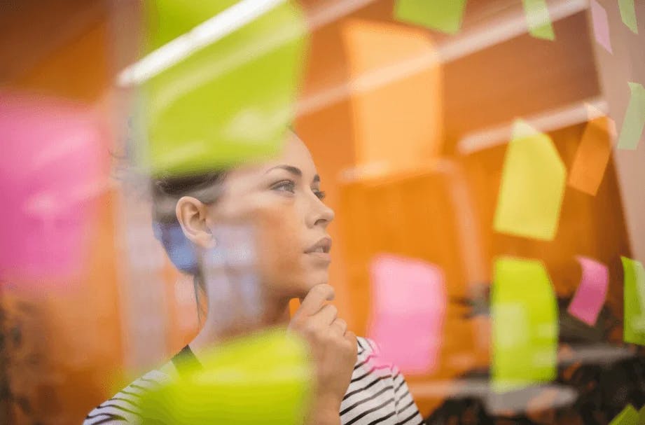 Mulher reflexiva analisando quadros de post-its coloridos em uma parede, focada em ideias criativas e planejamento colaborativo.