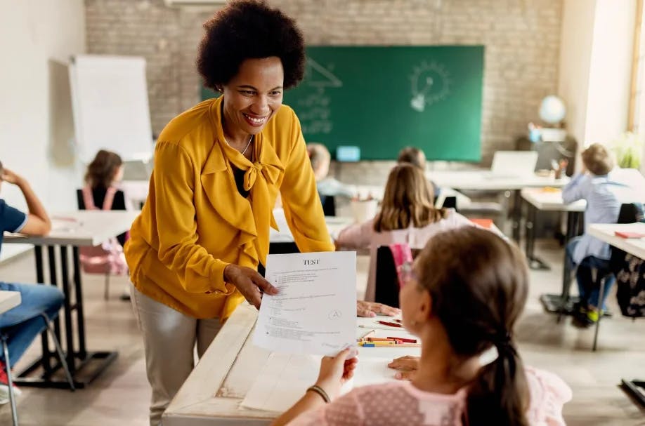Professora entregando um teste a uma aluna em sala de aula de forma sorridente e amigável, com alunos estudando ao fundo, promovendo educação e avaliação escolar.