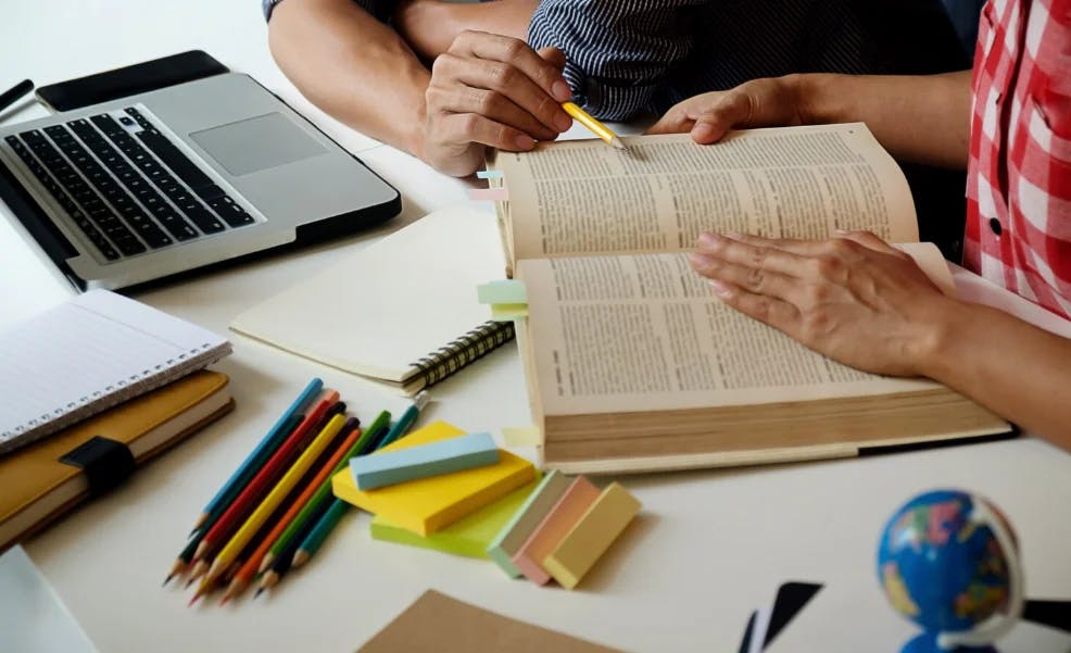 Grupo de estudantes estudando com livros, canetas coloridas e um laptop em uma mesa de estudos, em ambiente de aprendizado colaborativo.