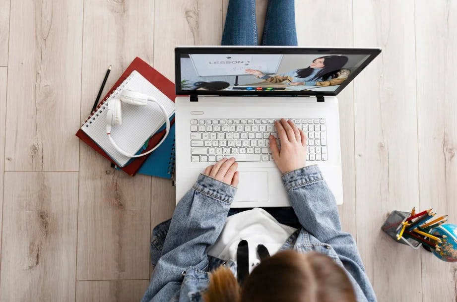 Estudante assistindo aula online em laptop, com materiais escolares ao redor, em ambiente doméstico, promovendo educação a distância.