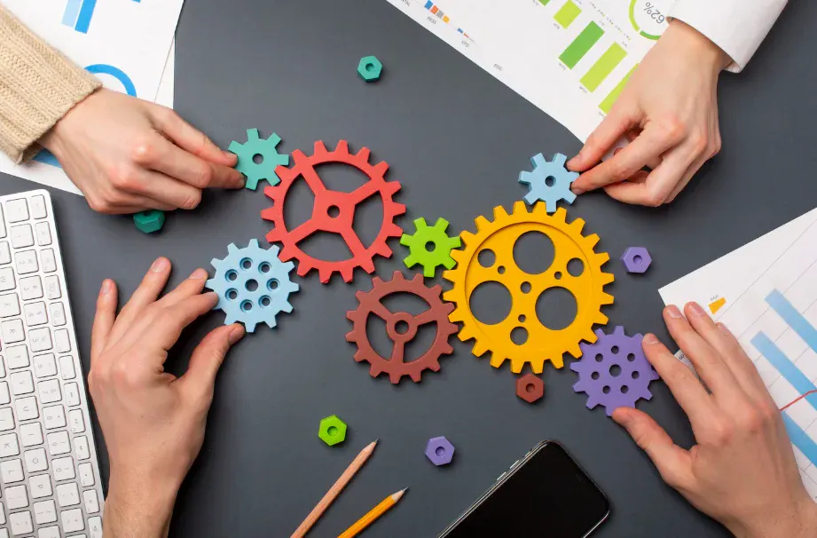 Pessoas analisando e ajustando engrenagens coloridas de diferentes tamanhos em uma mesa de trabalho para otimizar processos.