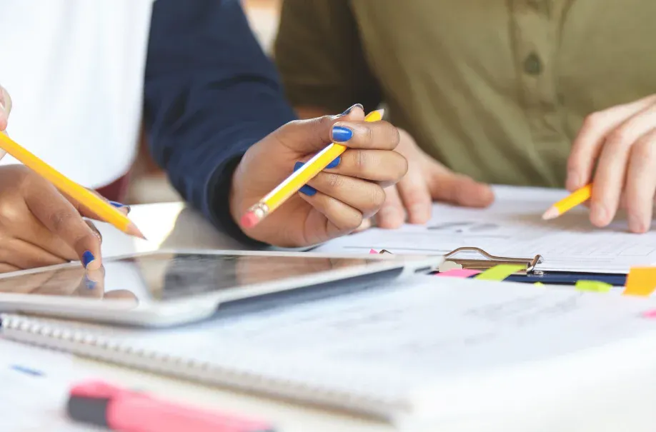 Pessoas trabalhando em uma reunião, utilizando lápis, tablets, cadernos e organizadores de papel para planejar e colaborar em um projeto de equipe.