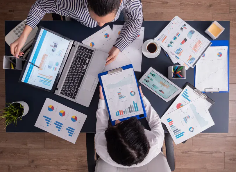 Pessoa analisando gráficos e relatórios de negócios em uma reunião de equipe com computadores, tablet e papéis com dados gráficos.