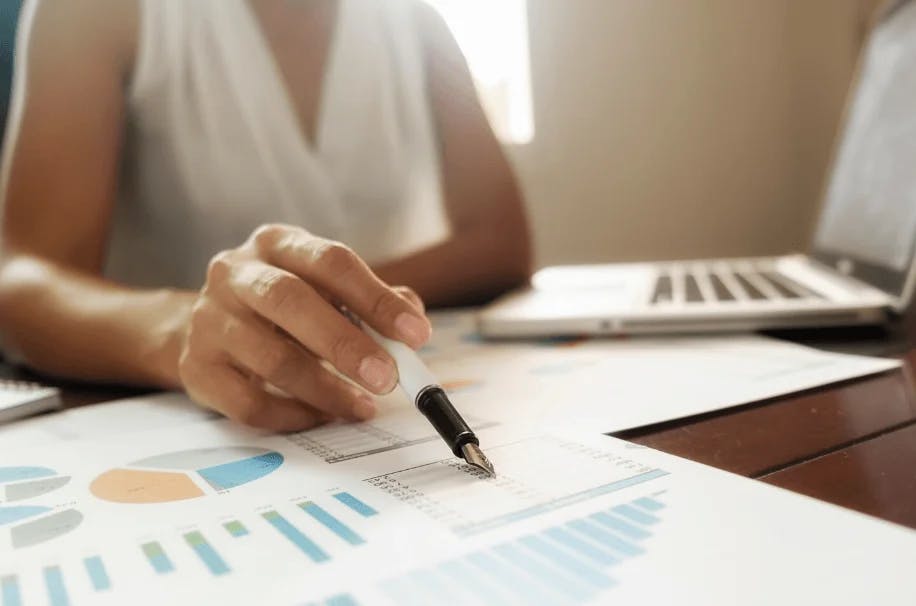 Pessoa analisando gráficos e dados financeiros em uma mesa de trabalho com laptop ao fundo, focada em estratégia de negócios e análise de mercado.