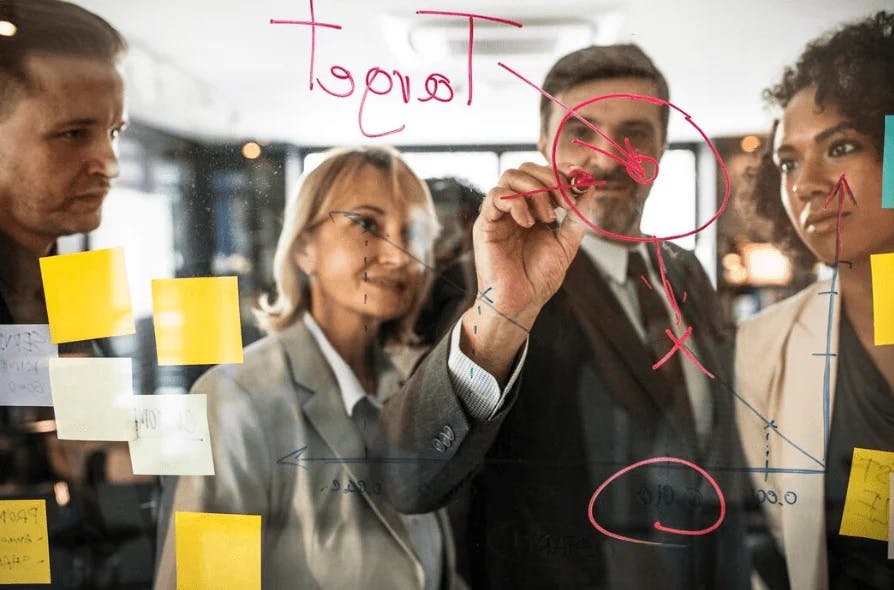 Equipe de profissionais colaborando e planejando estratégias em reunião de negócios, usando quadro branco com anotações e post-its para discussão.