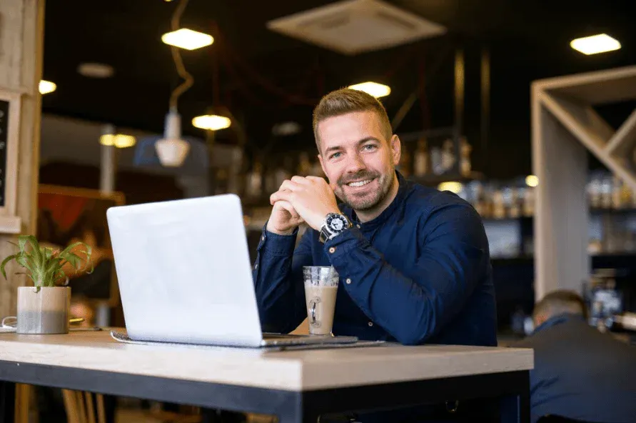 Homem sorridente usando laptop em cafeteria moderna, com bebida e plantas, ambiente aconchegante e descontraído.