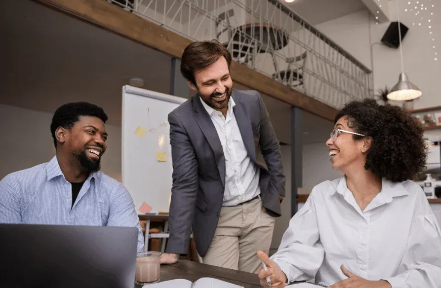 Três profissionais em uma reunião de trabalho, sorrindo e conversando em um ambiente de escritório moderno, promovendo colaboração e inovação.