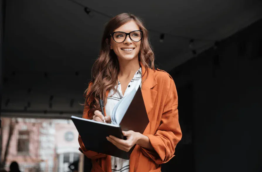 Mulher empresária sorridente com óculos pretos, vestindo blazer alaranjado e camisa branca, segurando um caderno e uma caneta, em ambiente urbano.