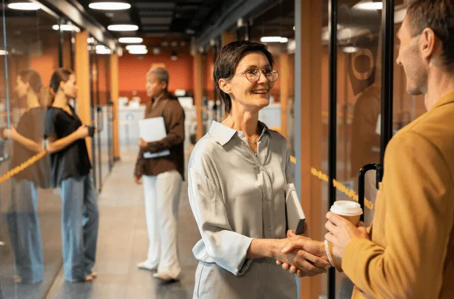 Mulher de óculos e cabelo curto apertando a mão de um homem sorridente em ambiente de espera. Fundo com outras pessoas na fila.
