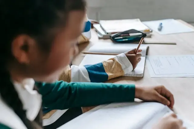 Criança estudando e fazendo tarefas escolares em uma mesa de estudos em casa, com caderno, livros e materiais escolares ao redor, focada na aprendizagem.