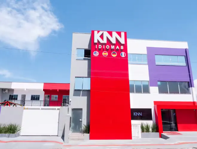 Imagem da fachada da loja KNN Idiomas, com destaque para o letreiro e fachada moderna em cores vermelha, branca e lilás, sob céu azul.