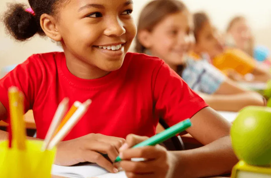 Criança sorrindo em sala de aula, ao lado de outros estudantes, ideal para conteúdo de educação infantil e aprendizagem escolar.