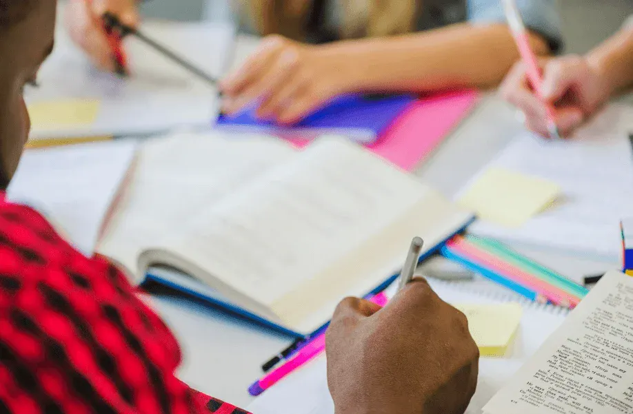 Reunião de estudantes ou profissionais trabalhando com anotações, cadernos e materiais de escritório em uma mesa de estudos ou reunião.