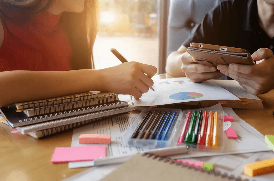 Duas pessoas estudando e trabalhando com materiais escolares e dispositivos eletrônicos em uma mesa de estudos, com foco em produtividade e organização.