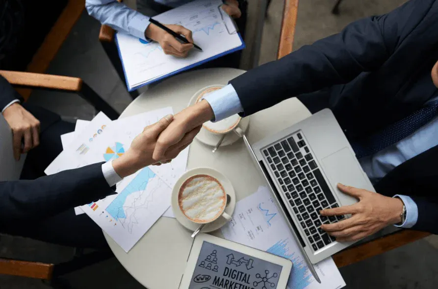 Negócios e marketing digital em reunião de profissionais, com pessoas apertando as mãos, usando laptops, gráficos e documentos relacionados ao marketing digital.