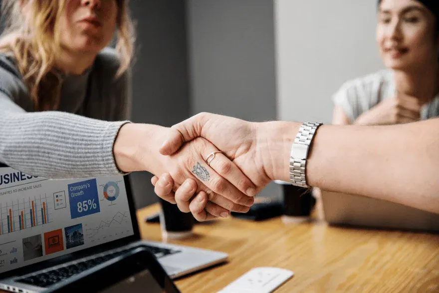 Imagem de duas pessoas apertando as mãos em um ambiente de negócios, símbolo de parceria e acordo empresarial, com uma mulher e uma jovem no fundo.