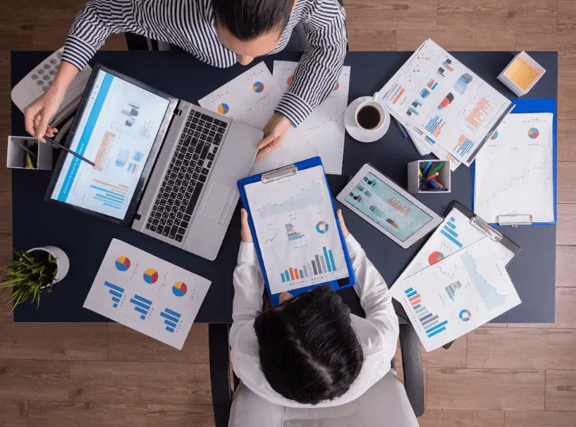 Imagem de duas pessoas analisando gráficos e dados em laptops, tablets e papéis, em um ambiente de escritório, com foco em análise de dados e apresentações.