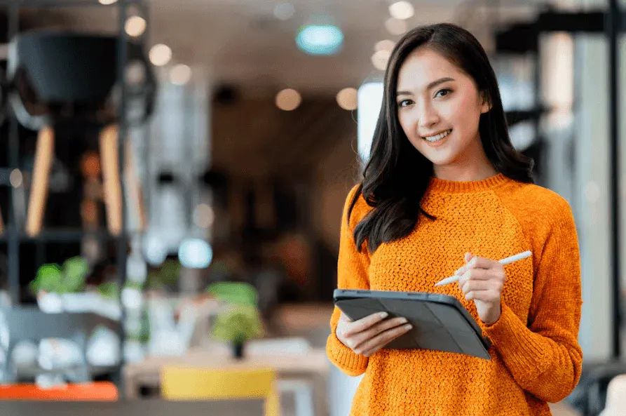 Mulher jovem sorridente usando suéter laranja e segurando tablet com caneta, no ambiente moderno de um café ou espaço de coworking.