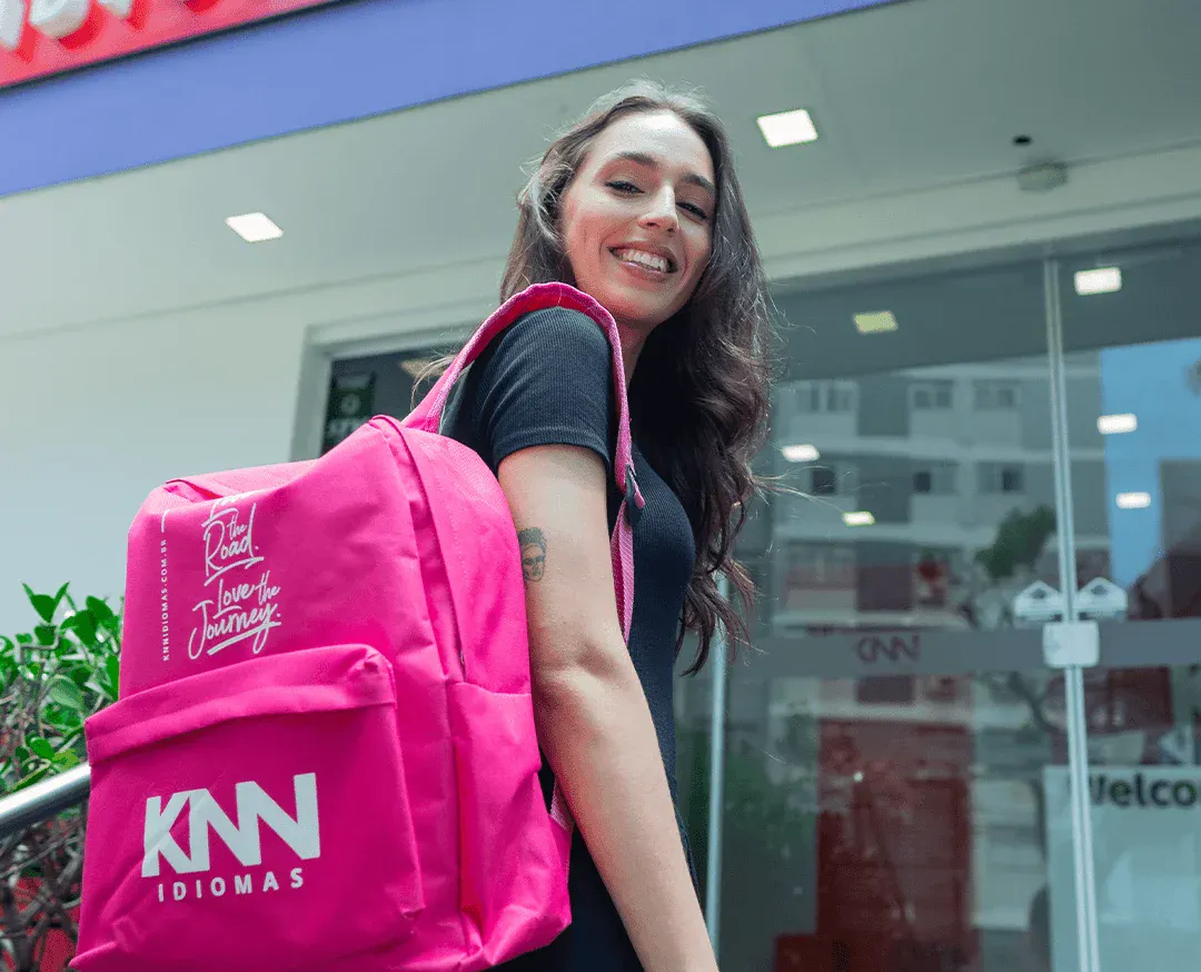 Mulher sorridente com mochila rosa na entrada de prédio de vidro, destacando a marca 'KNN Idiomas' e a frase 'I love the journey' na mochila.