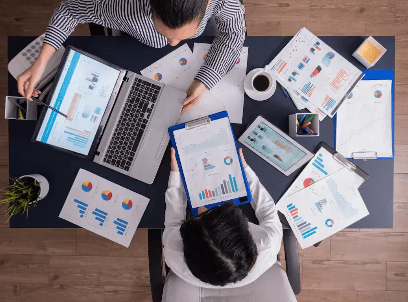 Duas pessoas trabalhando em uma mesa com computadores, tablets, papéis e gráficos, em ambiente de escritório, focadas em análise de dados e relatórios.