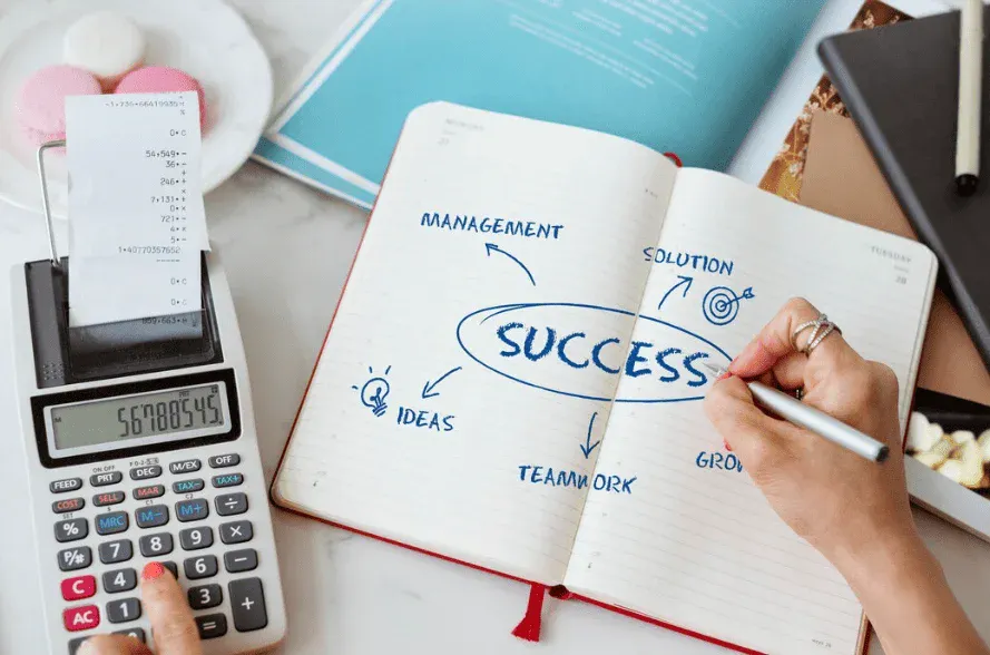 Pessoa escrevendo em caderno com palavras relacionadas ao sucesso, como gestão, soluções, ideias, equipe e crescimento, com materiais de escritório ao redor.