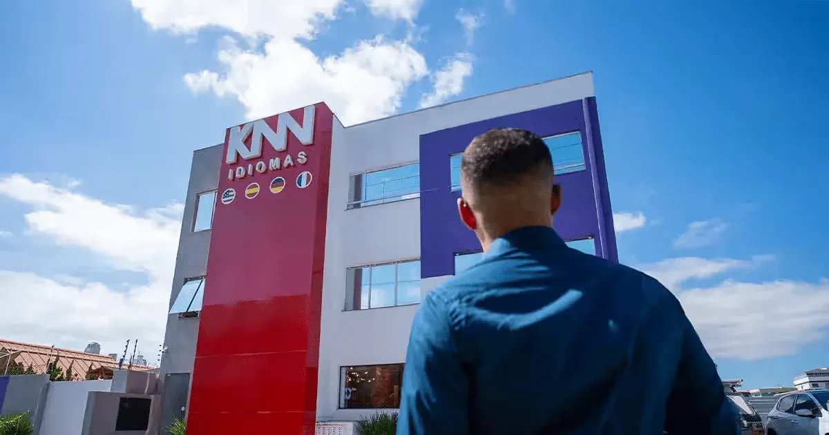 Pessoa observando o edifício da KNN Idiomas em uma tarde ensolarada, com céu azul e nuvens dispersas ao fundo.